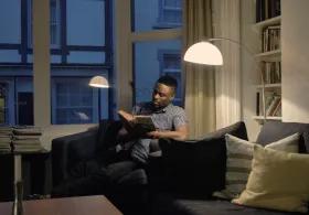 Man reading on a sofa in a dimly lit apartment, illuminated by a single overhead light