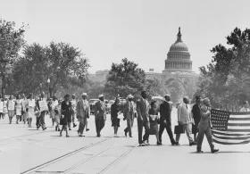 A black and white image of people marching in a protest, one holding an American flag, with trees and buildings in the background