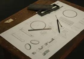 A table with engineering diagrams on paper, showing a compass, ruler, and pen, alongside an open brown case with its lid resting at an angle