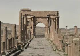 Stone-paved Roman road lined with weathered columns leading to a large, arched gate in the ruins of an ancient city, with rolling hills in the background.