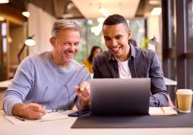 Two people smiling around a laptop with notes and coffee