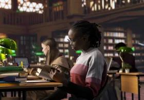 Students reading and working at desks in a quiet library setting, surrounded by shelves of books