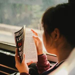 Back view of a woman sitting on a bus reading a book, with the open pages of the book visible and the bus interior softly blurred in the background