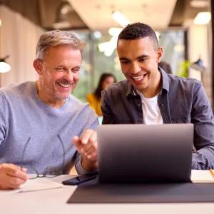 Two people smiling around a laptop with notes and coffee
