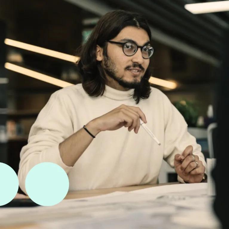 Man with shoulder length dark hair, sat at desk with pencil in hand