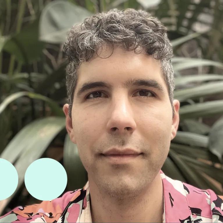 Man with greying dark curly hair, stood in front of large plant