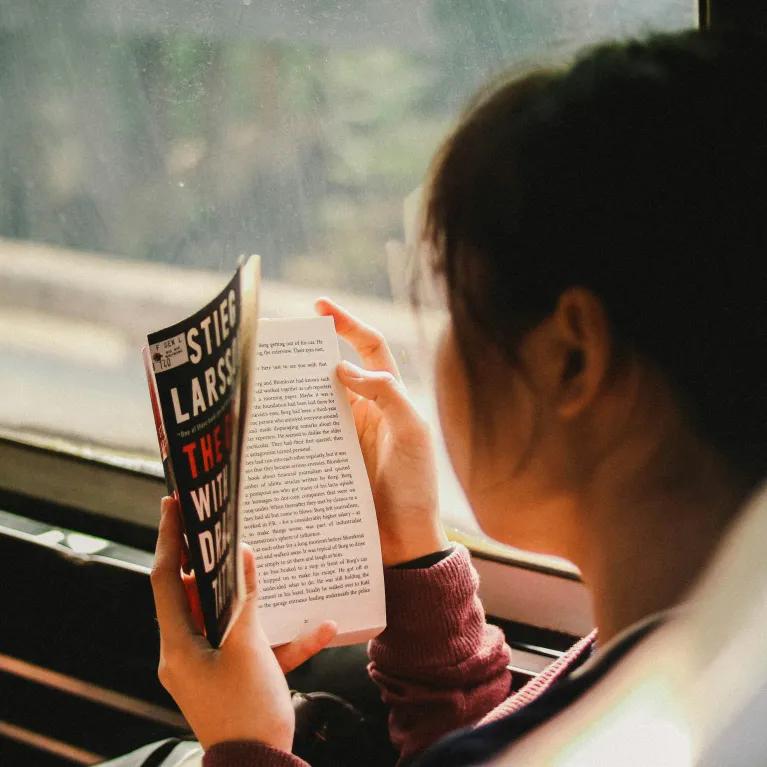Back view of a woman sitting on a bus reading a book, with the open pages of the book visible and the bus interior softly blurred in the background