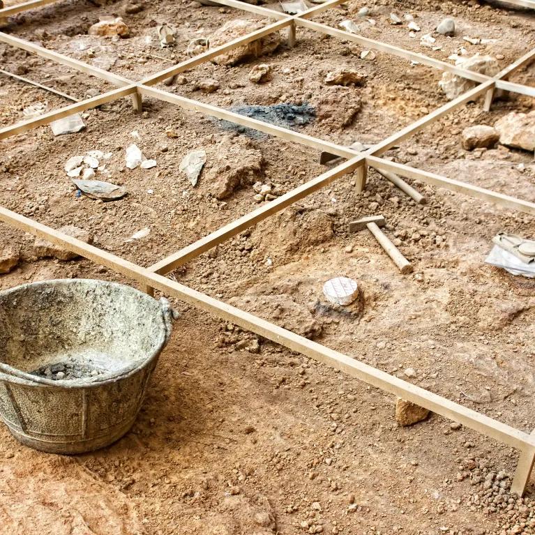 An archaeological excavation site with fossils exposed beneath a wooden grid, surrounded by a bucket and excavation tools on the dirt