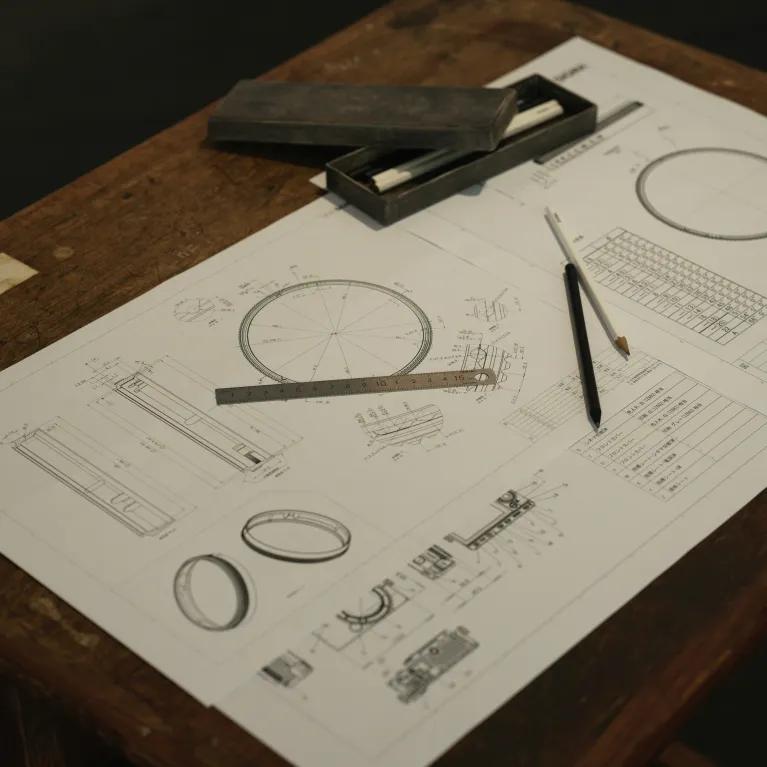 A table with engineering diagrams on paper, showing a compass, ruler, and pen, alongside an open brown case with its lid resting at an angle