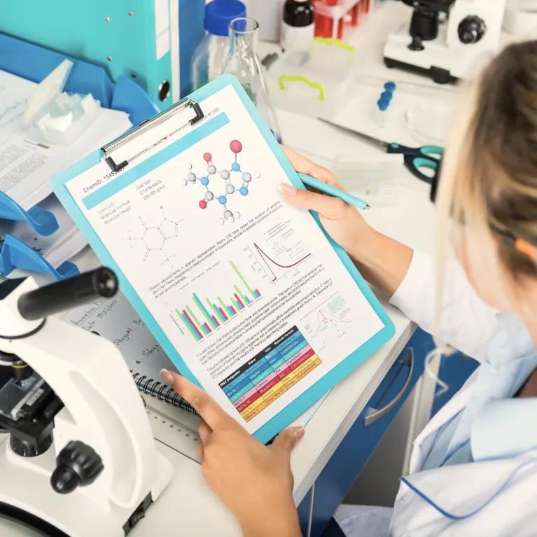 Person in lab coat looking at notes with microscope on lab bench