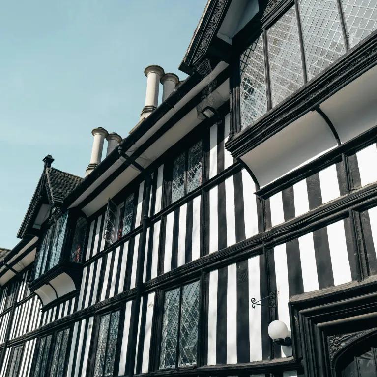 Timber-framed Tudor-style building with leaded glass windows and tall chimneys, viewed from below against a clear sky