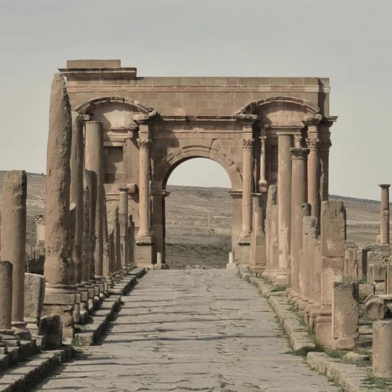 Stone-paved Roman road lined with weathered columns leading to a large, arched gate in the ruins of an ancient city, with rolling hills in the background.