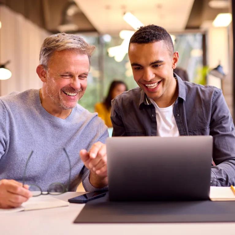 Two people smiling around a laptop with notes and coffee